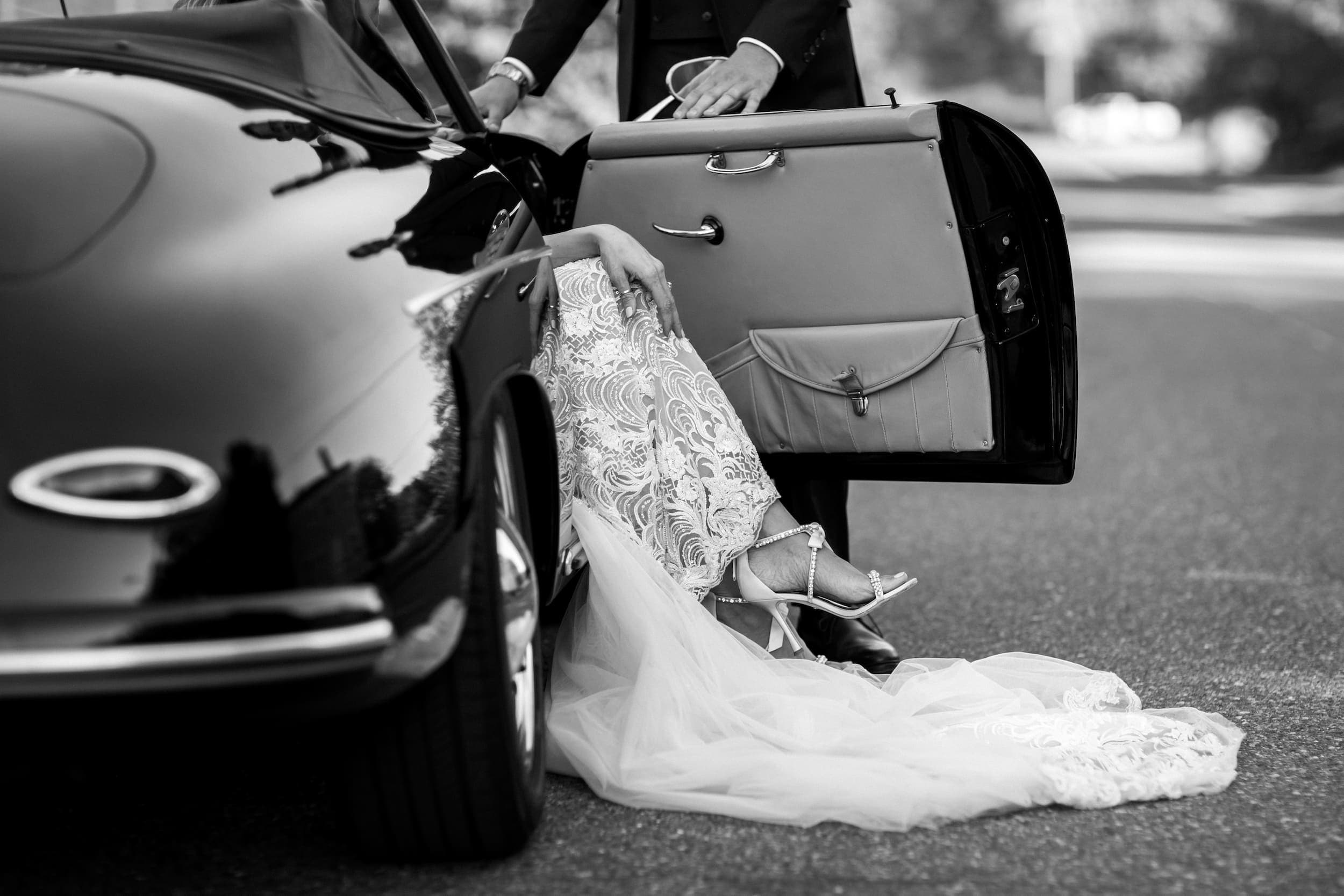 A black and white photo of a brides legs emerging from an old vintage black car on her wedding day