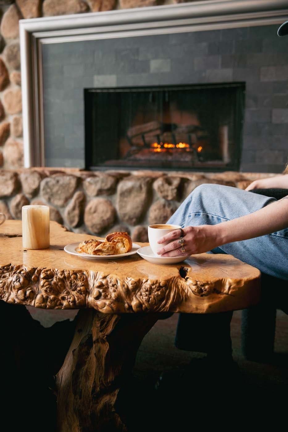 A fresh pastry and hot beverage on a live edge wood table in front of the fireplace at Edgewater
