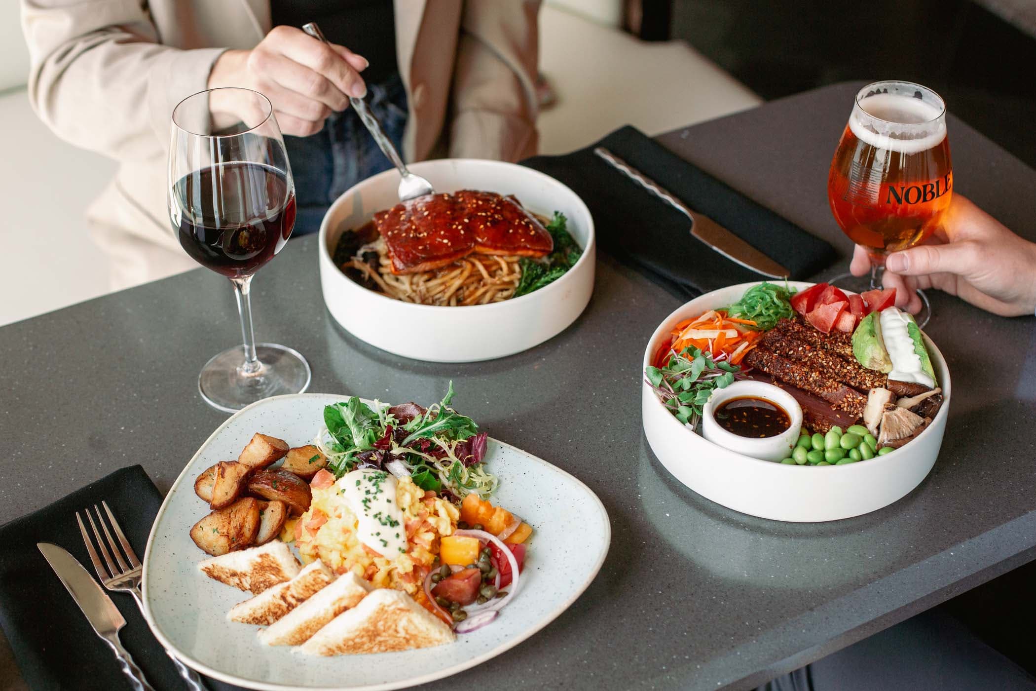 3 colorful dishes, a glass of beer, and red wine on a table inside Six Seven at Edgewater