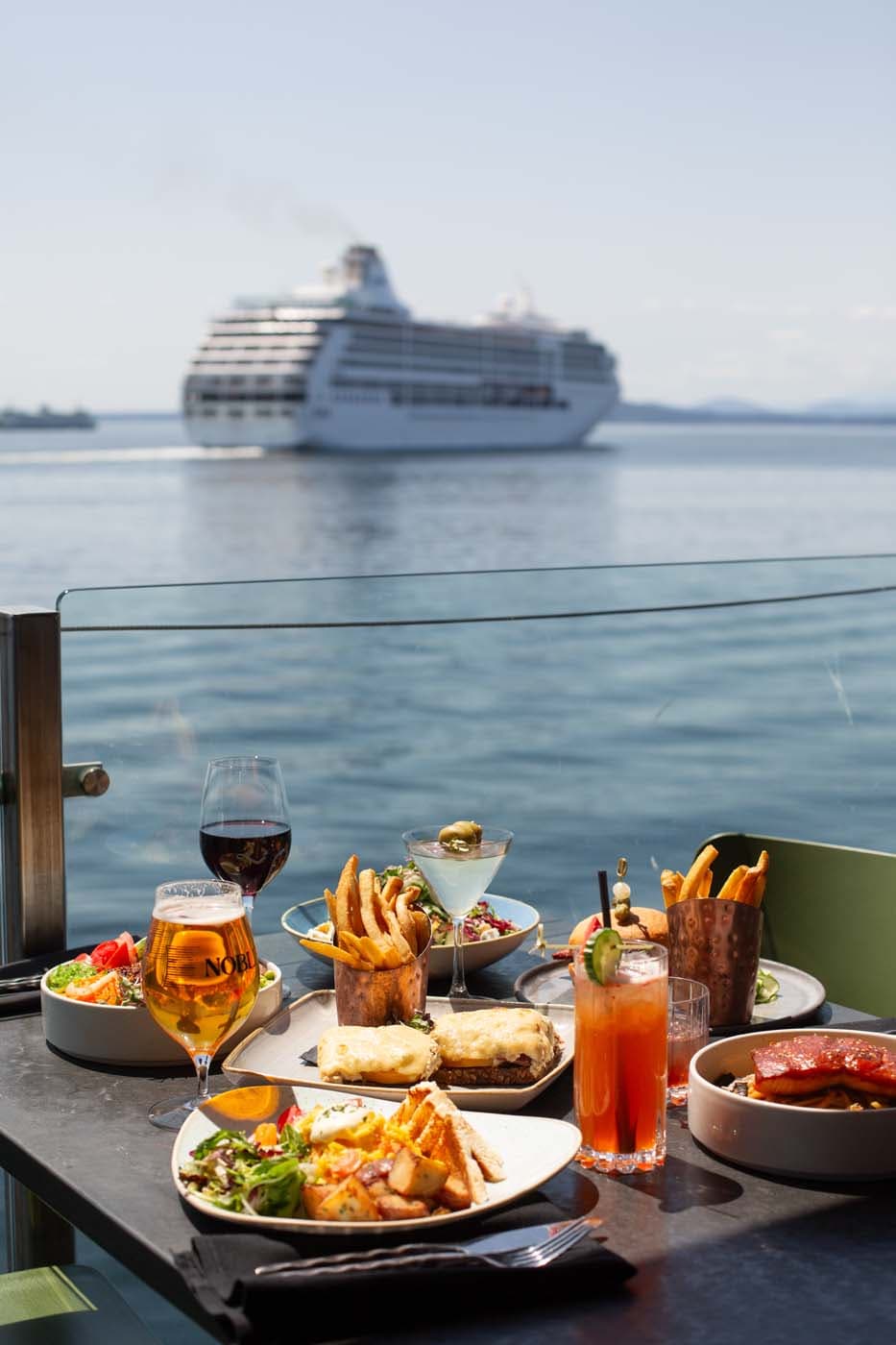 A variety of dishes on a. patio table at Edgewater with a cruise ship in the background