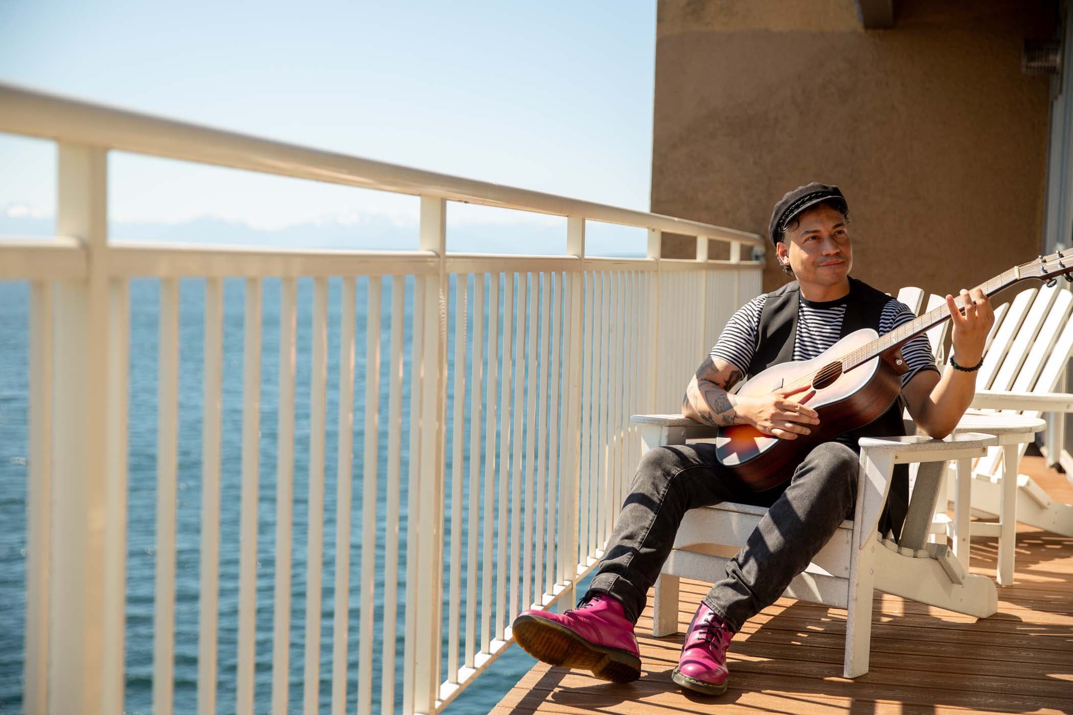 A man playing guitar and smiling on a room balcony over looking the water at Edgewater