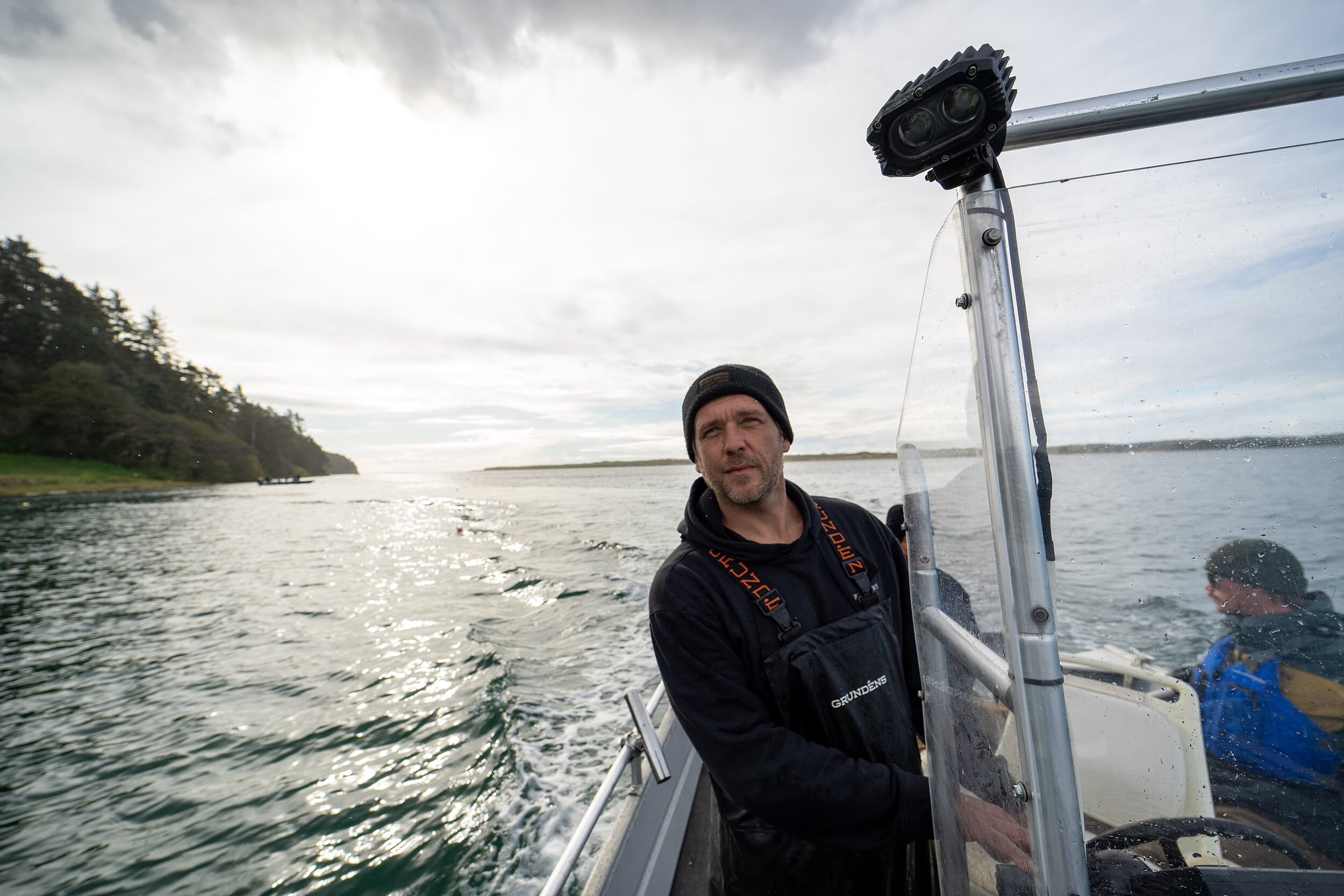 Fisherman in waders piloting a boat across calm Puget Sound waters under a moody Pacific Northwest sky, sourcing fresh local seafood for The Edgewater Hotel in Seattle